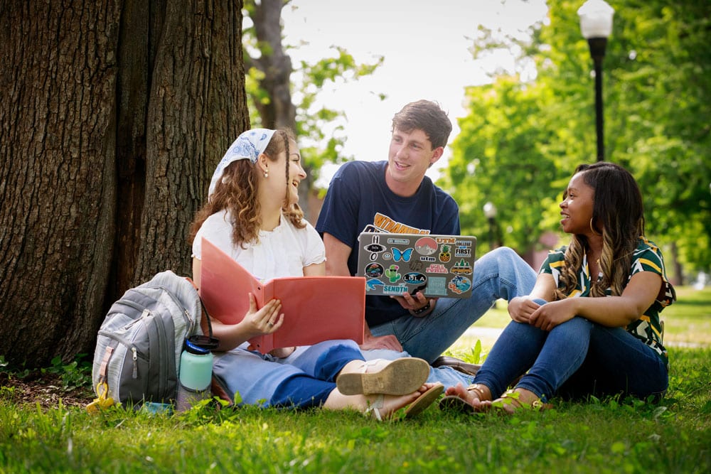 Three students sitting on grass, one holding binder and another holding laptop