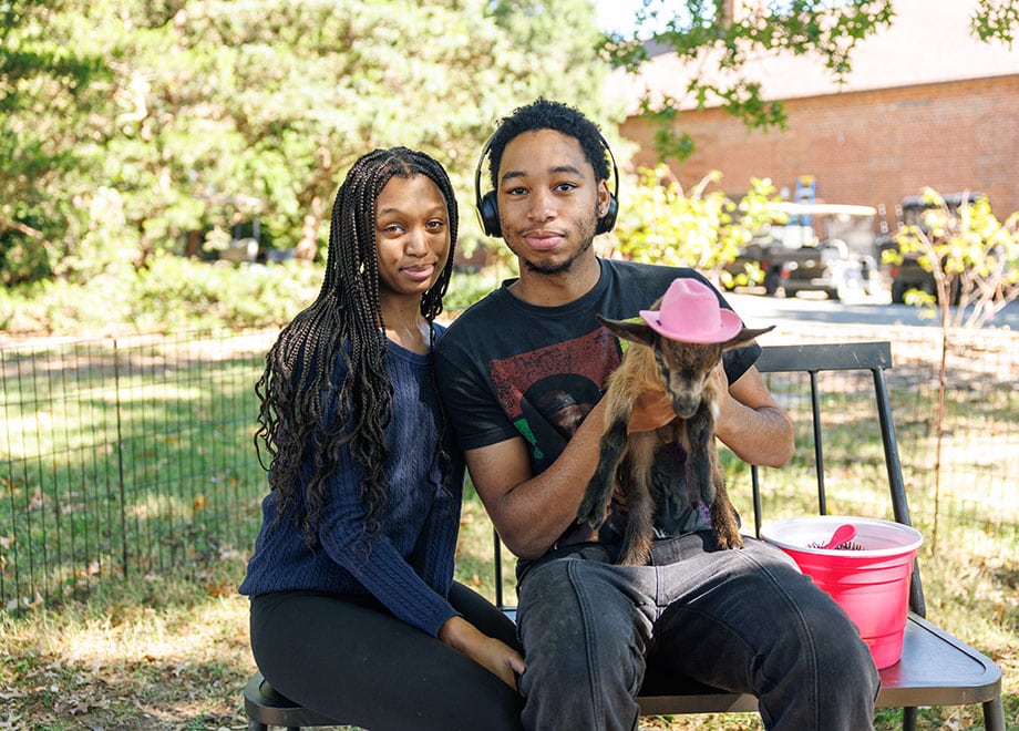 Two students hold a goat. The goat is wearing a pink hat.